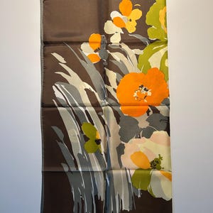 May include: A rectangular brown silk scarf with a floral design. The scarf features large orange, yellow, and green flowers with gray and white foliage. The edges are a darker brown.