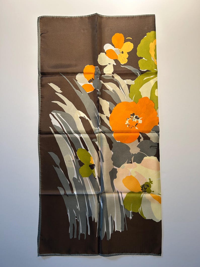 May include: A rectangular brown silk scarf with a floral design. The scarf features large orange, yellow, and green flowers with gray and white foliage. The edges are a darker brown.