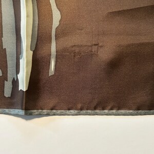 May include: A brown silk scarf with a geometric design in shades of gray and white. The scarf has a textured appearance and a gray border. The design features vertical and abstract shapes.
