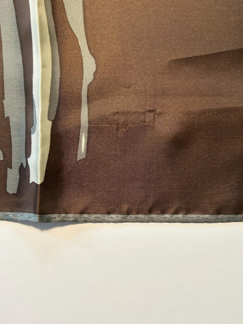 May include: A brown silk scarf with a geometric design in shades of gray and white. The scarf has a textured appearance and a gray border. The design features vertical and abstract shapes.
