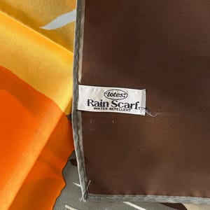 May include: A colorful scarf with a brown border and a label that reads "totes! Rain Scarf. WATER REPELLENT." The scarf features shades of orange, yellow, and gray.