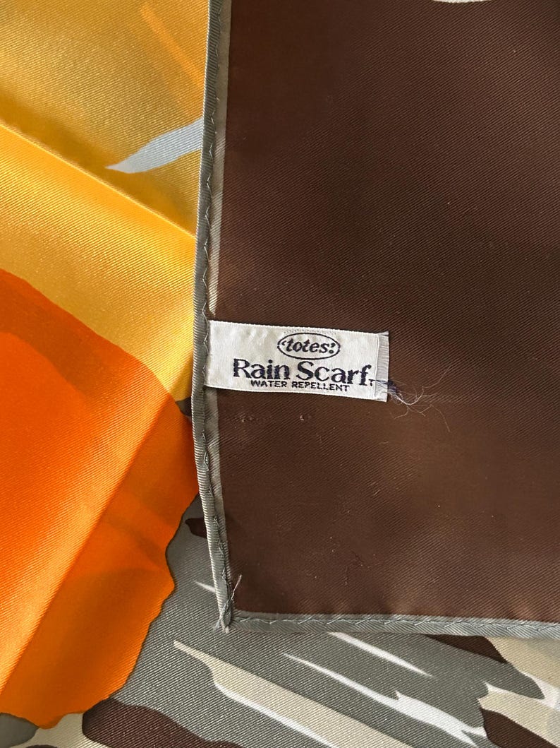 May include: A colorful scarf with a brown border and a label that reads "totes! Rain Scarf. WATER REPELLENT." The scarf features shades of orange, yellow, and gray.