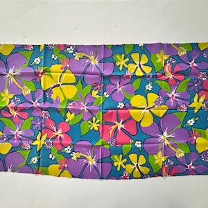May include: A rectangular fabric piece featuring a vibrant floral pattern with large purple, yellow, and pink hibiscus flowers set against a bright turquoise background, interspersed with small green leaves and tiny white blossoms across the surface.