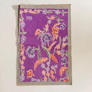 May include: A rectangular piece of purple fabric featuring a batik-style floral pattern in shades of orange, white, and dark purple. The design includes a decorative border of small flowers and the text MANFUT KRATON KIDOL printed in the upper left corner.