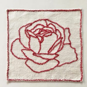 May include: A square off-white linen napkin featuring a hand-embroidered red rose design in the center. The rose is outlined with a decorative red stitch pattern, and the edges of the napkin are finished with a matching red stitched border.