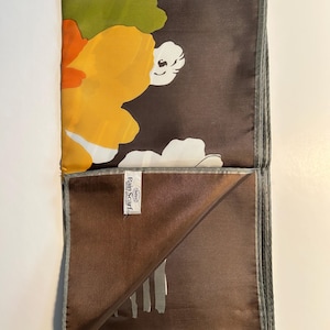 May include: A folded silk scarf with a floral pattern. The scarf features large yellow, orange, and white flowers against a dark brown background. The edges are trimmed with a gray border. The label reads "Rain Scarf".