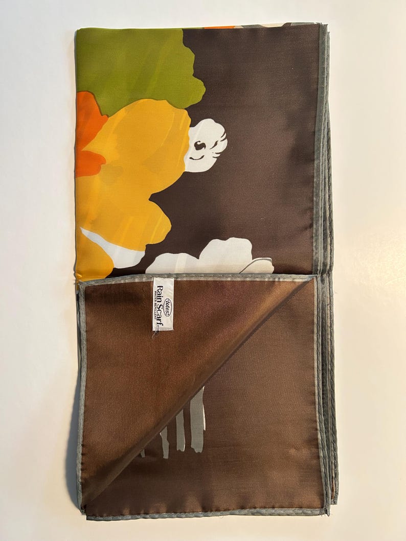 May include: A folded silk scarf with a floral pattern. The scarf features large yellow, orange, and white flowers against a dark brown background. The edges are trimmed with a gray border. The label reads "Rain Scarf".