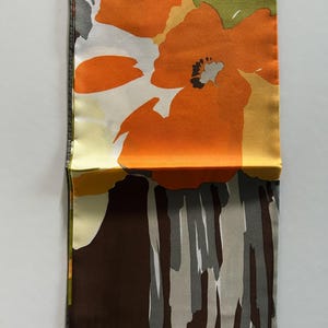 May include: Folded silk scarf with a floral pattern. The scarf features large orange flowers with black centers, set against a white and yellow background. The bottom portion has a brown and gray abstract design. The scarf is folded in half.