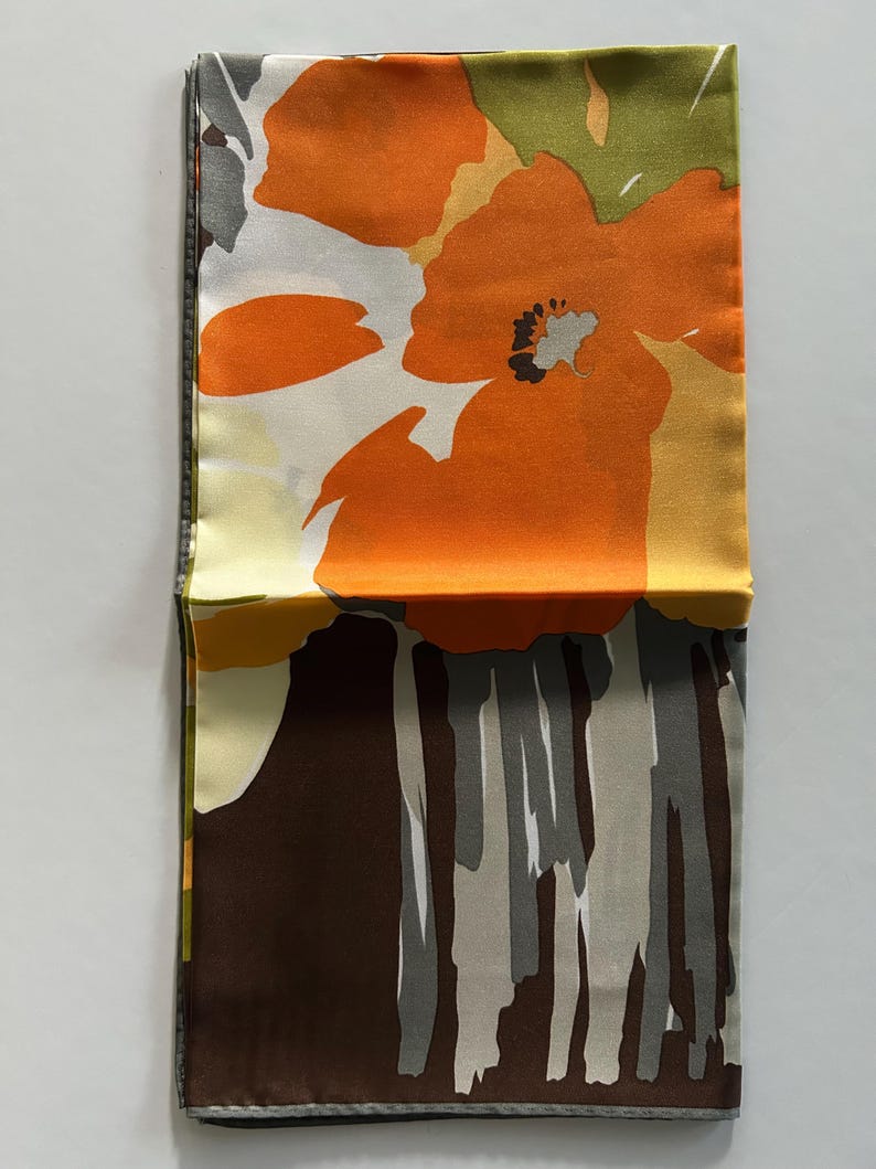 May include: Folded silk scarf with a floral pattern. The scarf features large orange flowers with black centers, set against a white and yellow background. The bottom portion has a brown and gray abstract design. The scarf is folded in half.