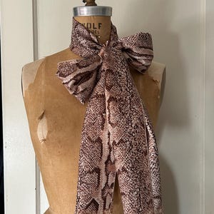 Puede incluir: Una bufanda con estampado de piel de serpiente marrón y beige está drapeada sobre un maniquí vintage. La bufanda está atada con un lazo en el cuello y tiene un borde con flecos. El maniquí es de color beige claro y tiene una parte superior de metal.