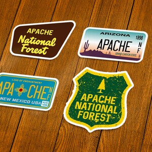 May include: Four stickers with designs related to the Apache National Forest. One sticker is a brown shape with "APACHE National Forest" in yellow text. Another is a license plate design with "APACHE" and "ARIZONA 1898". The third is a license plate design with "APACHE" and "NEW MEXICO USA". The fourth is a green shield with "APACHE NATIONAL FOREST".