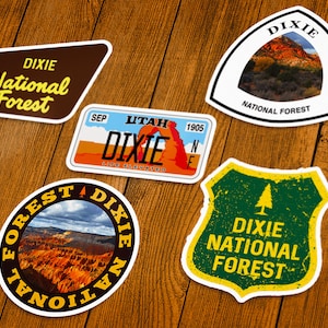 May include: A collection of Dixie National Forest stickers in various shapes. The stickers feature text and images of the forest, including a license plate design and a shield-shaped sticker with a pine tree.