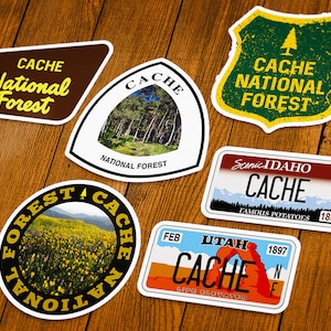 May include: A collection of Cache National Forest stickers in various shapes. Designs include a shield with a tree, a license plate with mountains, and a sticker with a field of yellow flowers. The stickers have the words "Cache" and "National Forest".
