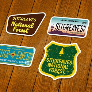 May include: Four Sitgreaves National Forest stickers. One is brown with yellow text, another is a license plate design with Arizona and the text "Sitgreaves". The third is a New Mexico license plate design. The last is a green shield with yellow text.
