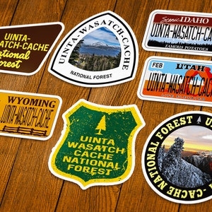 May include: A collection of Uinta-Wasatch-Cache National Forest stickers in various shapes. Designs include scenic views, license plate styles, and text such as "Wyoming" and "Idaho". The stickers are on a wooden surface.