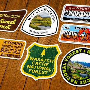May include: A collection of colorful stickers featuring the words "Wasatch-Cache National Forest" and other related text. The stickers have various shapes and designs, including a Wyoming license plate, a shield, and a circle. The stickers are on a wooden surface.