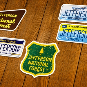 May include: A collection of stickers featuring the name "Jefferson" and "National Forest". Designs include license plates from Kentucky and Virginia, and a shield-shaped sticker with a tree graphic. The stickers are on a wooden surface.