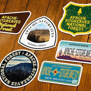 May include: A collection of seven stickers featuring the text "Apache-Sitgreaves National Forest." The stickers have various shapes, including a shield, a license plate, and a circle. Colors include brown, green, blue, and yellow.