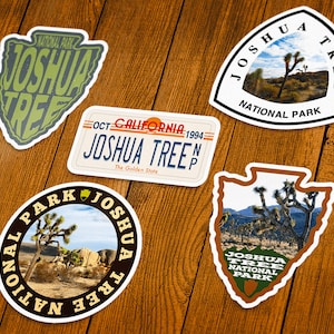 May include: A collection of Joshua Tree National Park stickers in various shapes. Designs include a license plate, a circular sticker with a desert landscape, and stickers with the text "Joshua Tree National Park".