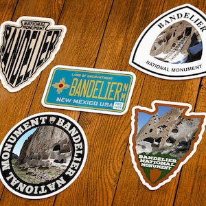 May include: A collection of Bandelier National Monument stickers on a wooden surface. The stickers feature various designs, including the monument's name, images of the landscape, and the New Mexico state flag.