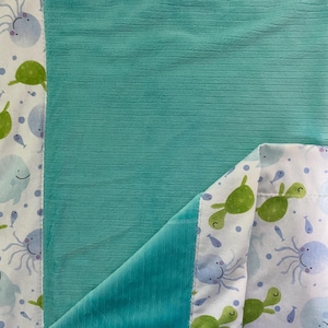 May include: A turquoise blue fleece blanket with a white fabric backing. The backing features a repeating pattern of green sea turtles, blue whales, and purple octopuses.
