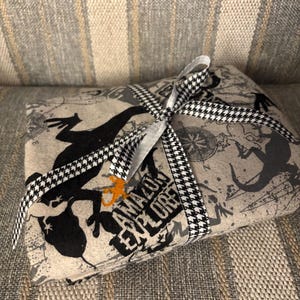 May include: A wrapped package with a black and white houndstooth ribbon. The package is wrapped in tan fabric with a map design and black silhouettes of frogs and the words "AMAZON EXPLORE".