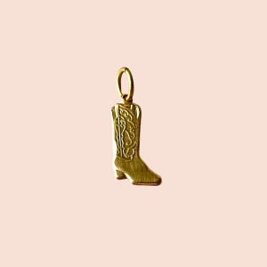May include: A gold-colored cowboy boot charm with intricate decorative detailing. The charm is suspended from a small loop, suggesting it's designed to be attached to a necklace or bracelet. The boot has a classic western design.