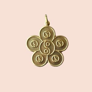 Può includere: Un ciondolo color oro a forma di fiore, con sei petali circolari. Ogni petalo presenta un simbolo stilizzato, con il petalo centrale che mostra un simbolo yin-yang. Il ciondolo ha un piccolo anello nella parte superiore per una catena.