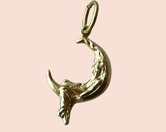 May include: A gold-colored pendant featuring a crescent moon with a figure seated within. The figure is draped in flowing garments. The pendant has a loop at the top for attaching to a chain. The overall design is detailed and ornate.