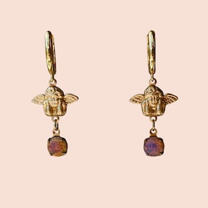 May include: A pair of gold-colored earrings. Each earring features a cherub pendant with a dangling, iridescent, square-cut gemstone. The earrings have a lever-back closure. The background is a solid, light peach color.