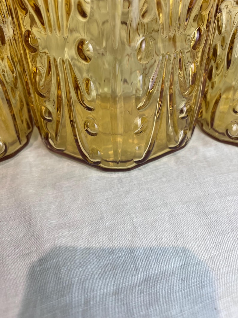 May include: Close-up of several amber-colored glass drinking glasses. The glasses have a textured, patterned design with rounded shapes and vertical lines. The glasses are on a white surface.