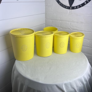 May include: A set of five yellow plastic containers with lids. Each container has a decorative floral design and is arranged on a round table covered with a white cloth. The containers vary in size.