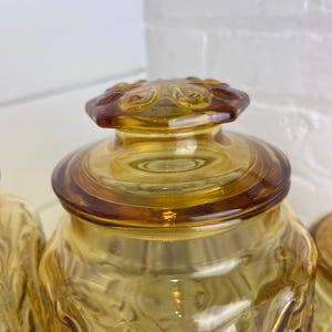 May include: Amber-colored glass jar with a decorative lid. The jar has a textured pattern on the sides and a round lid with a floral design. The glass has a warm, golden hue, and the jar is likely used for storage.