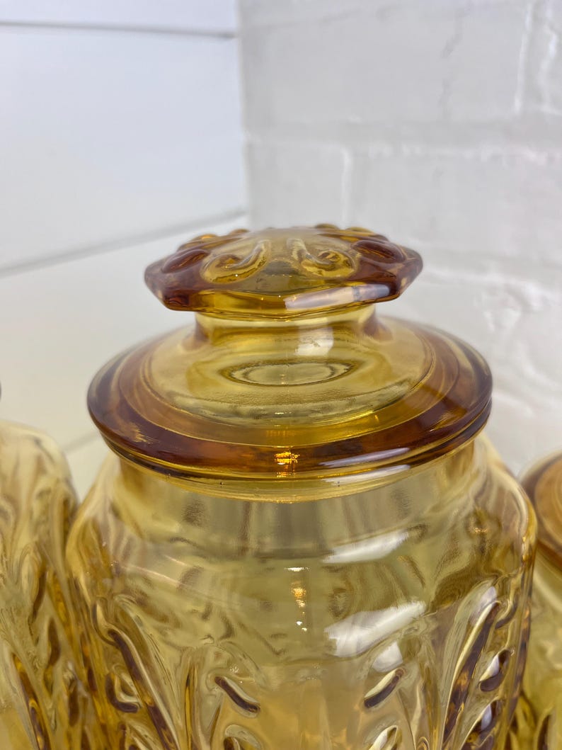 May include: Amber-colored glass jar with a decorative lid. The jar has a textured pattern on the sides and a round lid with a floral design. The glass has a warm, golden hue, and the jar is likely used for storage.