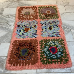 May include: A rectangular, hand-tufted rug with a salmon-colored background. The rug features six square designs with floral patterns in shades of brown, green, blue, and purple. The rug is made of a soft, textured material.