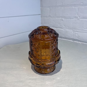 May include: Amber-colored glass candle holder with a decorative, textured pattern. The candle holder has a round base, a cylindrical body, and a domed lid. The glass has a warm, golden hue, and the design features geometric shapes.