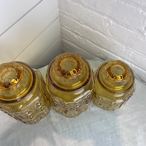 May include: Three amber-colored glass jars with lids. The jars have a decorative floral pattern on the sides and a round, textured lid. The jars are arranged in a row, with the lids facing up.