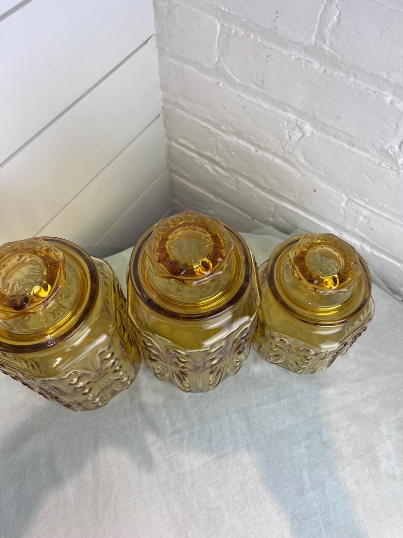 May include: Three amber-colored glass jars with lids. The jars have a decorative floral pattern on the sides and a round, textured lid. The jars are arranged in a row, with the lids facing up.