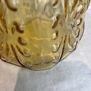 May include: Close-up of a vintage amber-colored glass pitcher. The pitcher has a textured, embossed design and a curved handle. The glass is transparent, allowing light to pass through, and sits on a white surface.