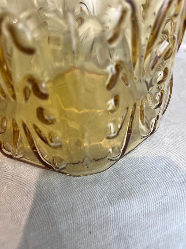 May include: Close-up of a vintage amber-colored glass pitcher. The pitcher has a textured, embossed design and a curved handle. The glass is transparent, allowing light to pass through, and sits on a white surface.