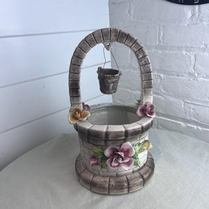 May include: A ceramic wishing well planter with a brick-like design. The well features a curved arch, a hanging bucket, and is adorned with pink and yellow floral accents. The planter sits on a round base and is suitable for indoor or outdoor use.