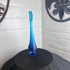 May include: A tall, slender, blue glass vase with a flared top and a hexagonal base. The vase is a vibrant shade of blue and stands on a black surface. The vase is likely used for decorative purposes.