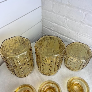 May include: A set of vintage amber-colored glass containers. The set includes three hexagonal jars with ornate embossed designs and three matching lids. The jars and lids are arranged on a white surface.