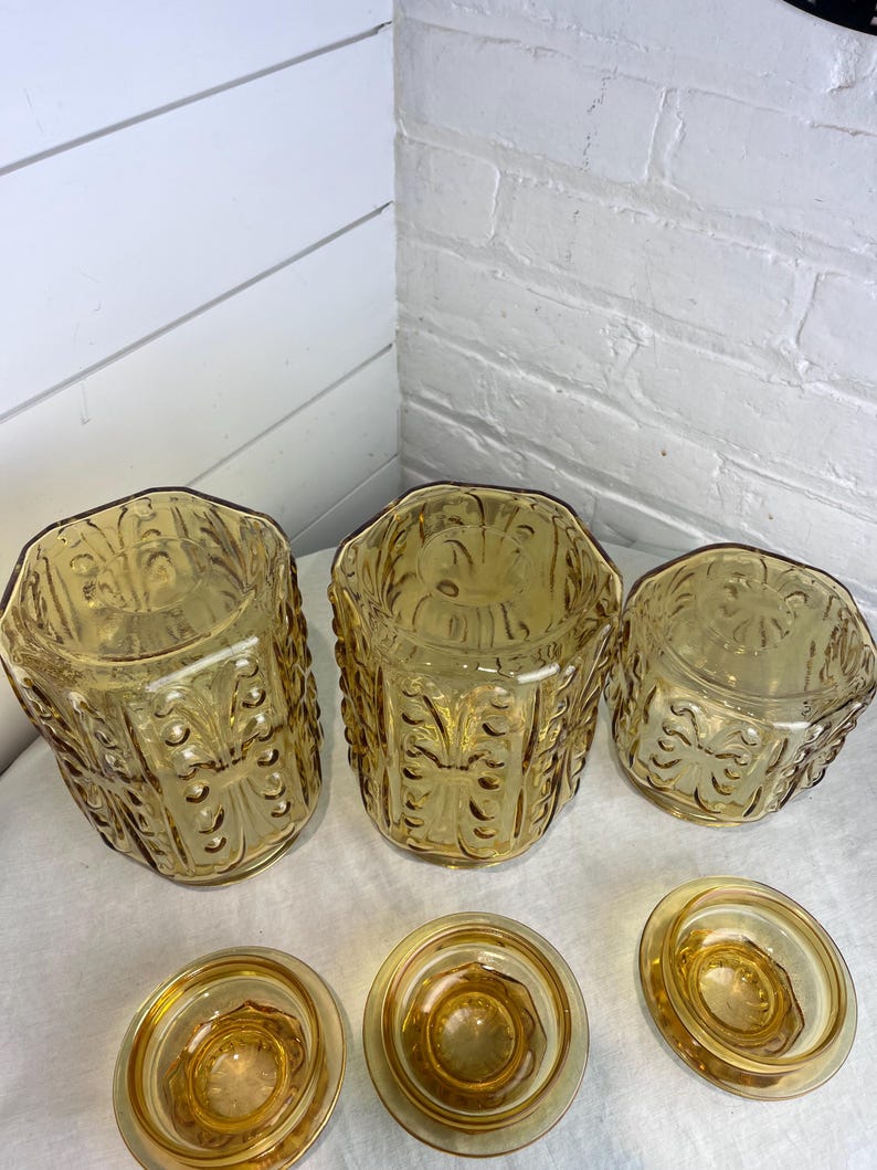 May include: A set of vintage amber-colored glass containers. The set includes three hexagonal jars with ornate embossed designs and three matching lids. The jars and lids are arranged on a white surface.