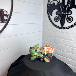 May include: A ceramic figurine featuring a green donkey pulling a cart with a large orange mushroom with white spots. A small figure sits on the mushroom. The donkey has brown hair and the cart has a brown wheel with yellow accents.