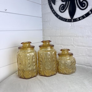 May include: Three amber-colored glass jars with lids. The jars have a decorative embossed pattern and are arranged in a row. The jars are of varying sizes, with the largest on the left and the smallest on the right.