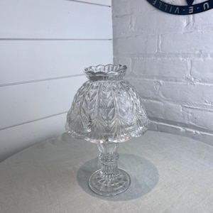 May include: A clear glass lamp shade featuring a scalloped top edge and a textured leaf pattern across the dome. The shade is supported by a matching clear glass pedestal base with a fluted stem and a circular foot, creating a cohesive piece.