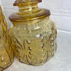 May include: A vintage amber glass jar with a lid. The jar has a decorative floral pattern and a round lid with a knob. The glass has a warm, golden hue. The jar is likely used for storage.