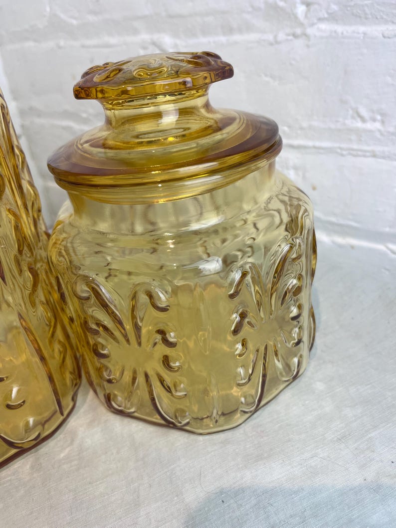 May include: A vintage amber glass jar with a lid. The jar has a decorative floral pattern and a round lid with a knob. The glass has a warm, golden hue. The jar is likely used for storage.
