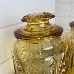 May include: Amber-colored glass jar with a decorative lid. The jar has a textured, embossed pattern and a matching lid with a floral design. The jar is likely used for storage and adds a vintage touch to any kitchen.